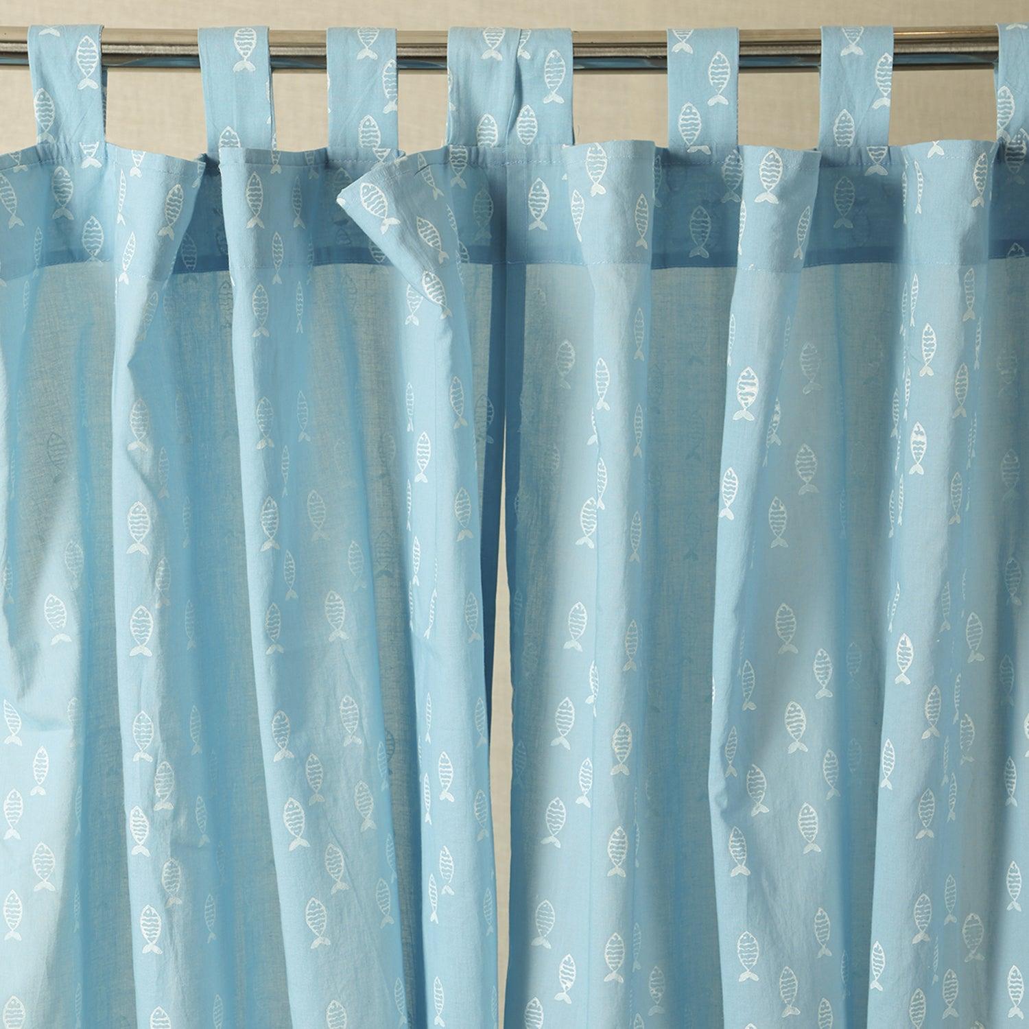Hand dyed blue mulmul cotton curtain with hand block printed fish motifs hanging by a window