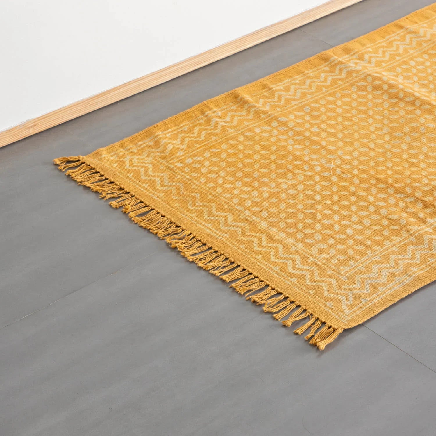 Oblique high-angle view of geometric handblock printed cotton rug in mustard and off-white, styled on floor