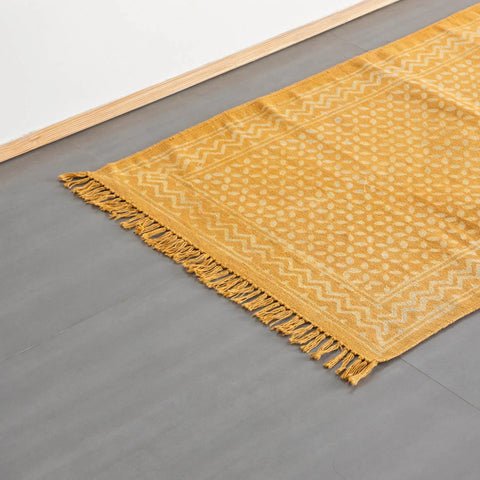 Oblique high-angle view of geometric handblock printed cotton rug in mustard and off-white, styled on floor