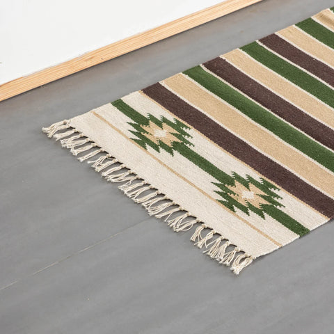 A flat weave runner rug with green, brown, and tan horizontal stripes and geometric motifs, featuring white-colored fringe on the floor.