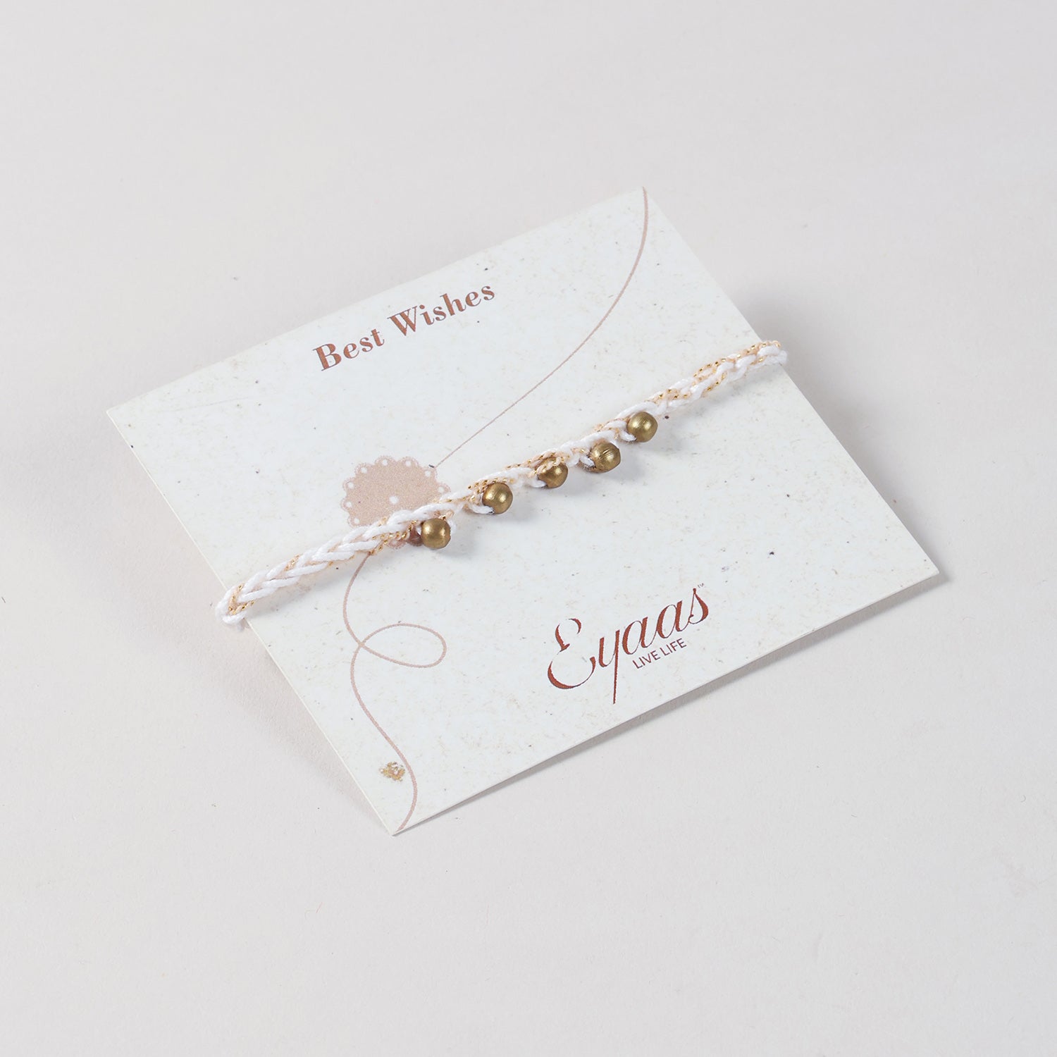 Angled view of the Handmade Crochet Brass Bead Rakhi with cream and gold threads attached to a festive Best Wishes card for Raksha Bandhan gifting. 