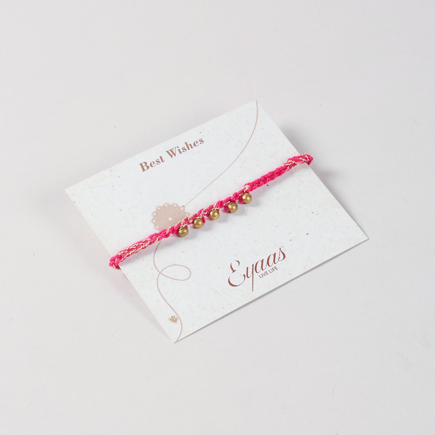Angled view of the Handmade Crochet Brass Bead Rakhi with pink and golden threads attached to a festive Best Wishes card for Raksha Bandhan gifting.