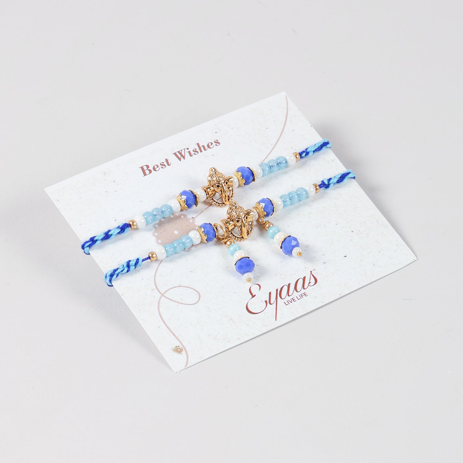 Angled view of the Handmade Ganesh Rakhi & Lumba Set with Blue Beads attached to a Best Wishes greeting card for Raksha Bandhan gifting.