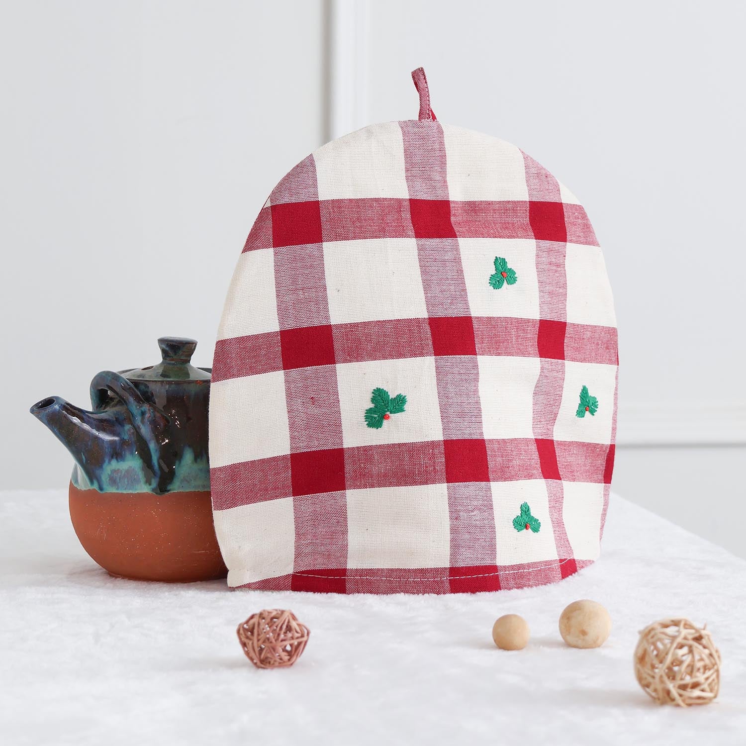 Christmas-themed cotton tea cozy with red and off-white check pattern placed over a teapot on a festive table setting with tea cups and holiday décor