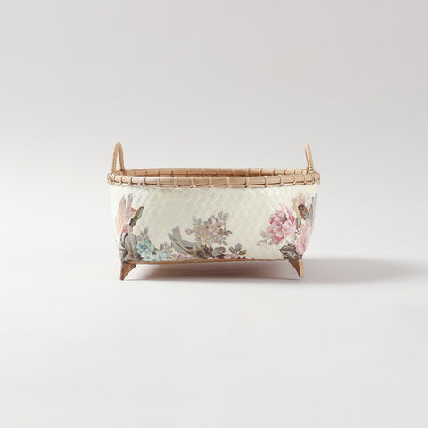 Side profile of a square wicker basket featuring pink floral decoupage art, sturdy side handles, and raised wooden feet