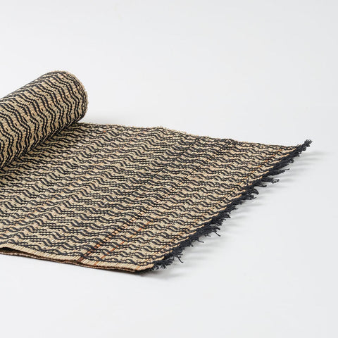 Angled view of a handmade Madurkathi table runner showcasing flexible natural fibre weaving and black tassel fringe 