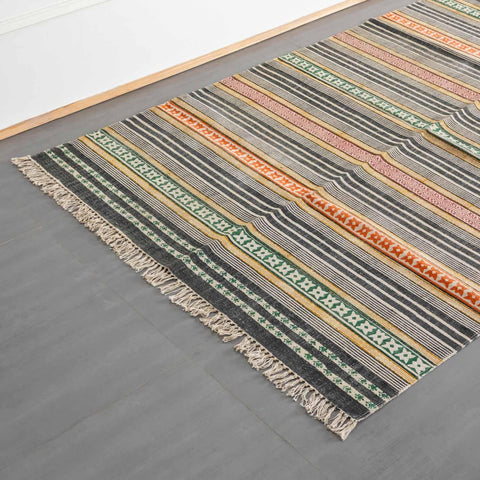 An oblique, high-angle view of a long, woven runner rug featuring multi-colored geometric stripes and cream fringe accents on a dark gray floor.