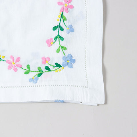 Detailed corner view of a handcrafted table runner featuring colourful floral vine embroidery along the border on a white cotton base