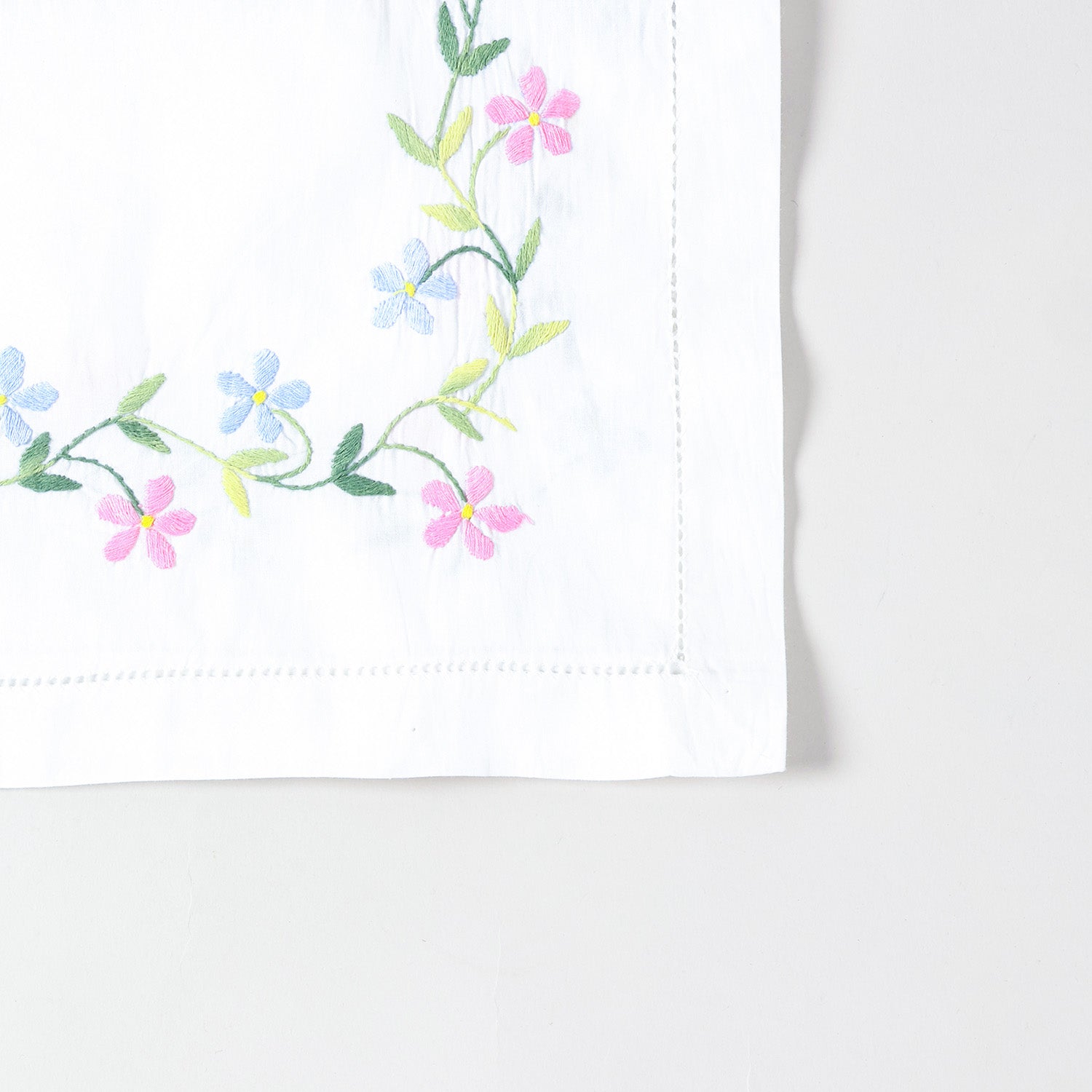 Detailed corner view of a handcrafted table runner featuring colourful daisy embroidery along the border on a white cotton base