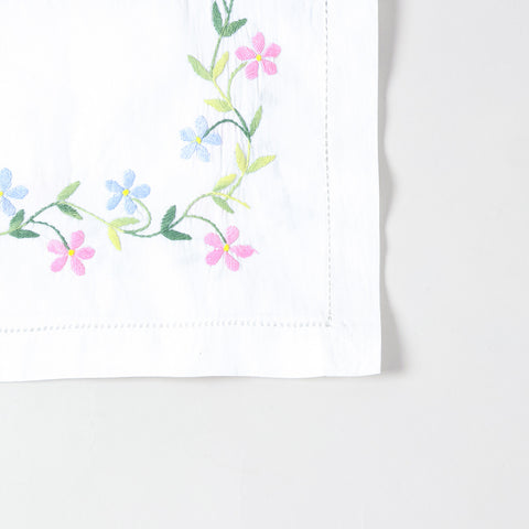 Detailed corner view of a handcrafted table runner featuring colourful daisy embroidery along the border on a white cotton base