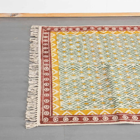 Top view of a mustard and red cotton dhurrie rug, highlighting geometric block prints and woven fringe detailing