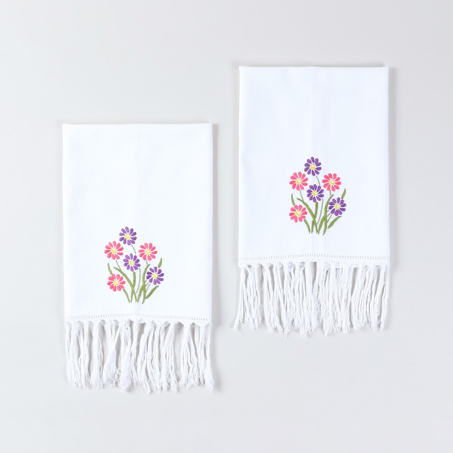 Set of two white cotton kitchen towels featuring pink and purple daisy floral embroidery and tasselled edges