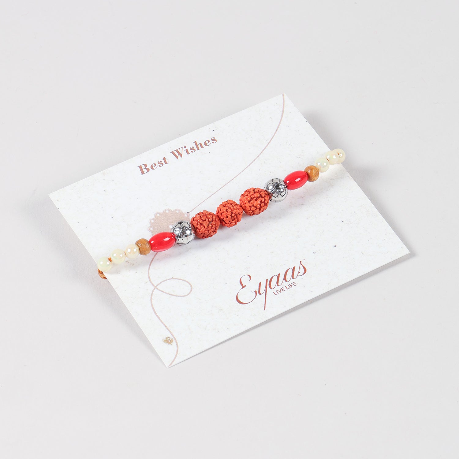 Angled view of the Handmade Rudraksha with Red and Pearl Bead Rakhi attached to a Best Wishes greeting card for Raksha Bandhan gifting.