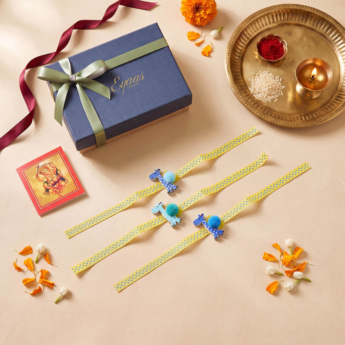 Set of 3 handcrafted wooden giraffe kids rakhis arranged with yellow ribbons, displayed next to a blue Eyaas gift box with a teal satin ribbon and a traditional brass puja thali for Raksha Bandhan gifting.