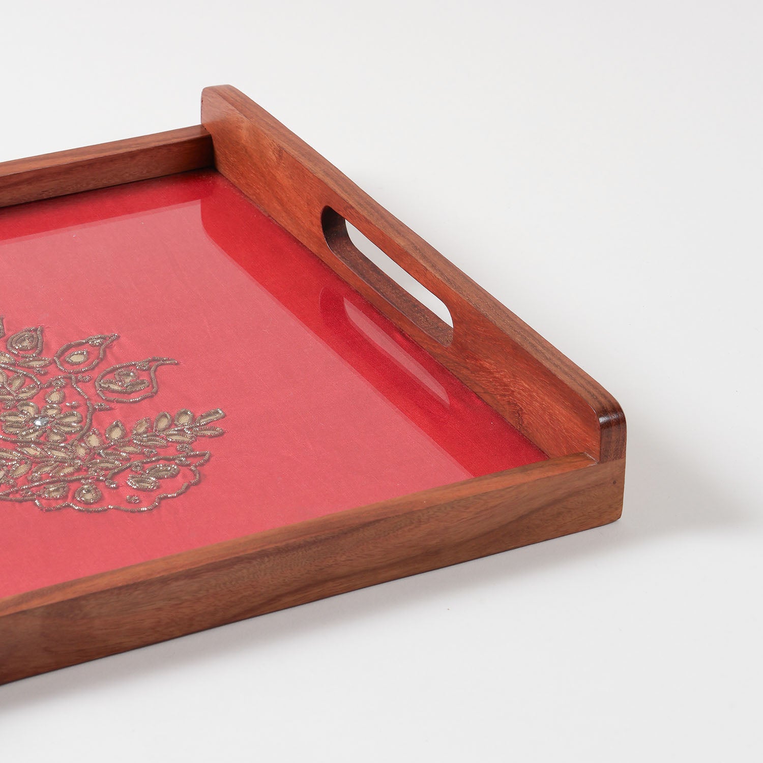 Corner angled close-up of a handcrafted rectangular sheesham wood serving tray with cutout handle, featuring red fabric with golden gota patti floral design under a glass top