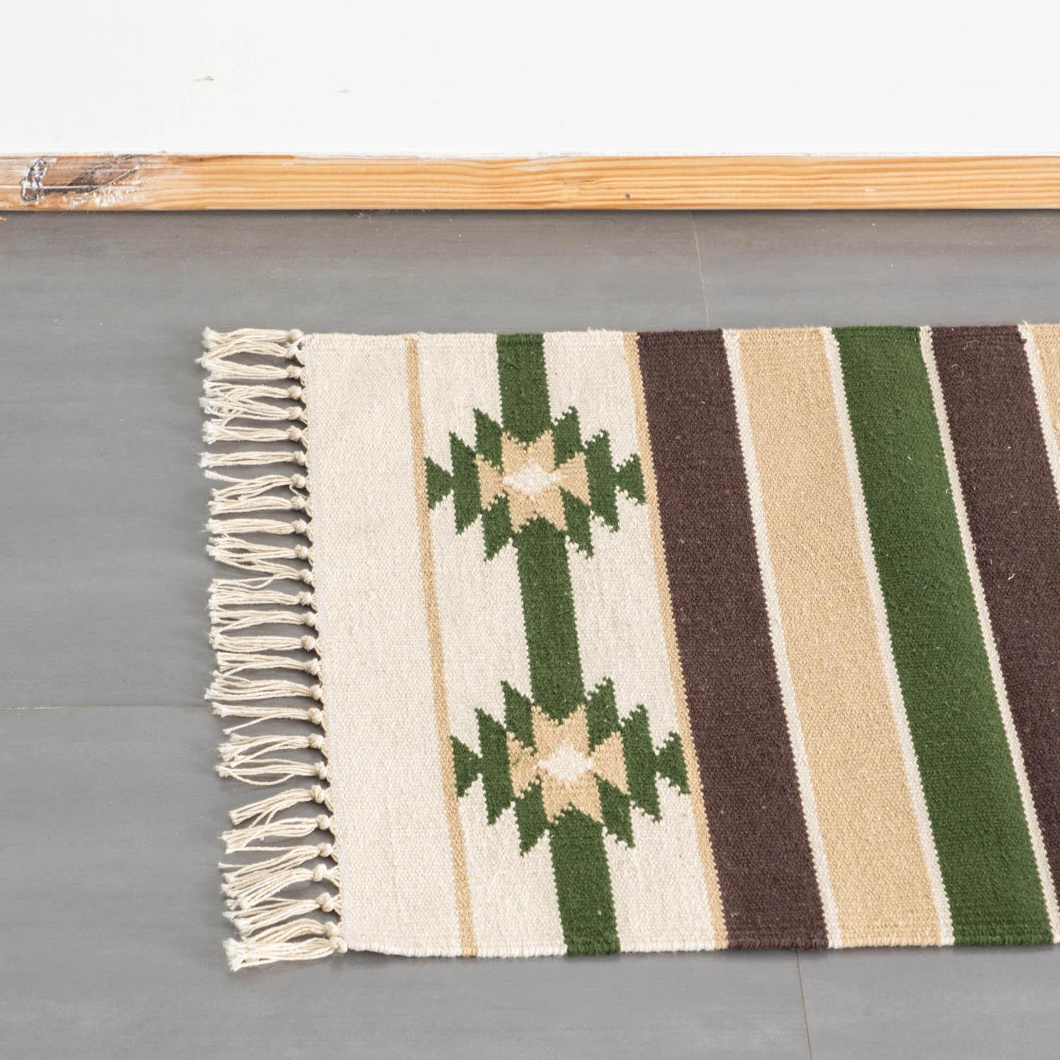 A high-angle top view of a part of a woven runner rug featuring geometric star motifs alongside green, brown, and white stripes, finished with a knotted white fringe on the left.