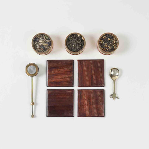 Top view of a tea gift hamper contents featuring tea blends, tea brewer, brass spoon, and handcrafted wooden coasters